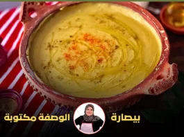 : طبق بيسارة في وعاء فخاري أحمر محاط بأوعية توابل وزيت الزيتون على مفرش مخطط باللونين الأحمر والأبيض.