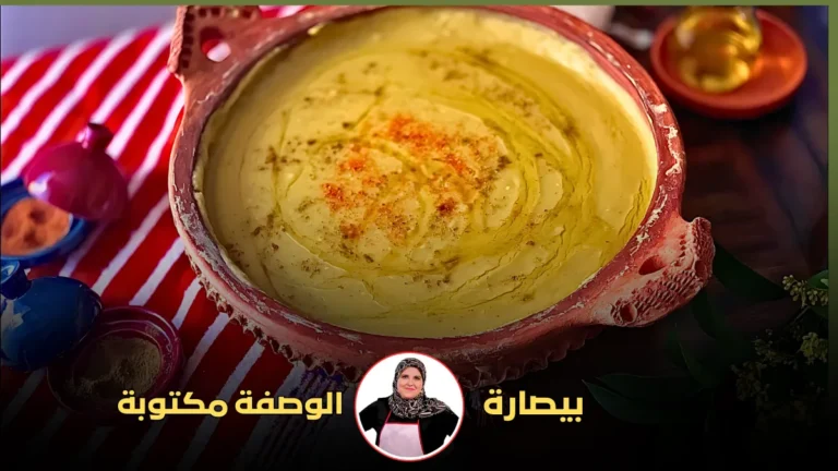 : طبق بيسارة في وعاء فخاري أحمر محاط بأوعية توابل وزيت الزيتون على مفرش مخطط باللونين الأحمر والأبيض.