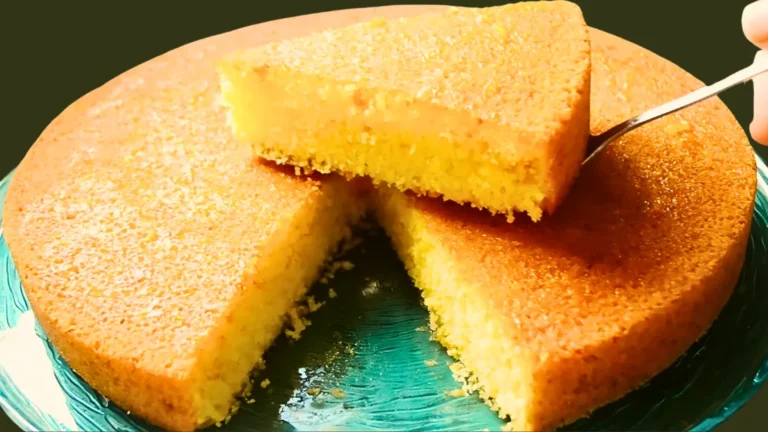 صورة تُظهر كيكة سميد دائرية بلونها الذهبي المميز، مع قطعة مرفوعة تُبرز قوامها الطري والهش. وضعت على طبق زجاجي أزرق أنيق.